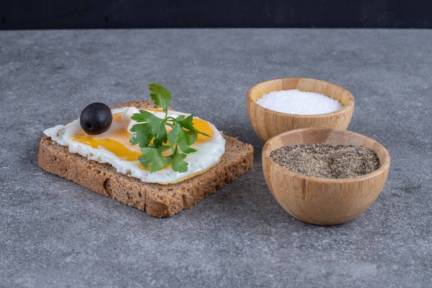 A slice of brown bread topped with a fried egg, parsley, and an olive, accompanied by bowls of salt and pepper on a gray surface.