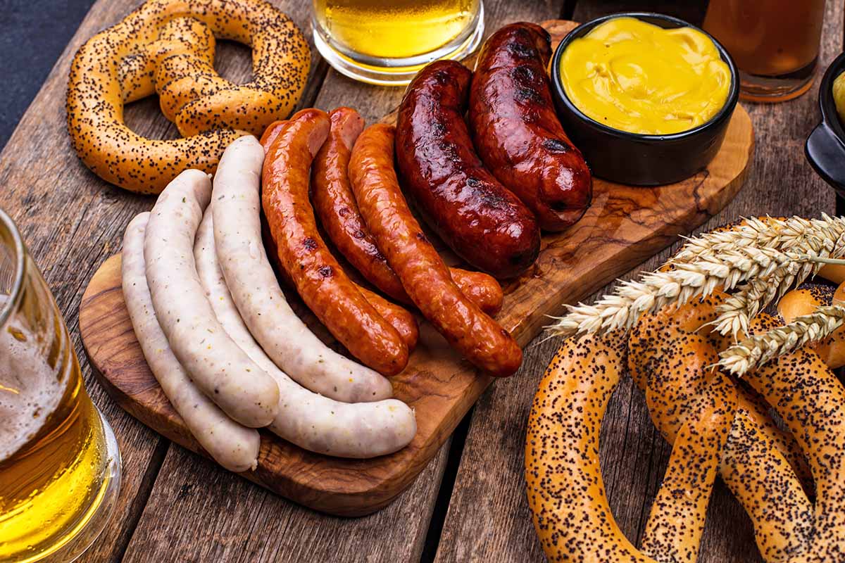 A wooden platter of assorted sausages, mustard, and pretzels with beer glasses in a rustic setting.