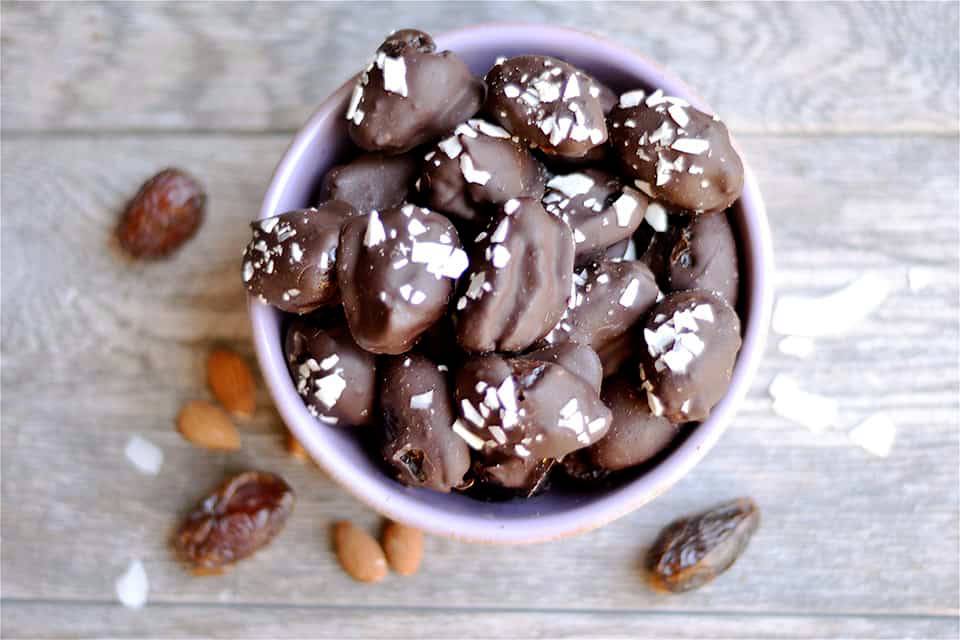 A bowl filled with chocolate-covered nuts and dates, garnished with coconut flakes, on a rustic wooden surface.