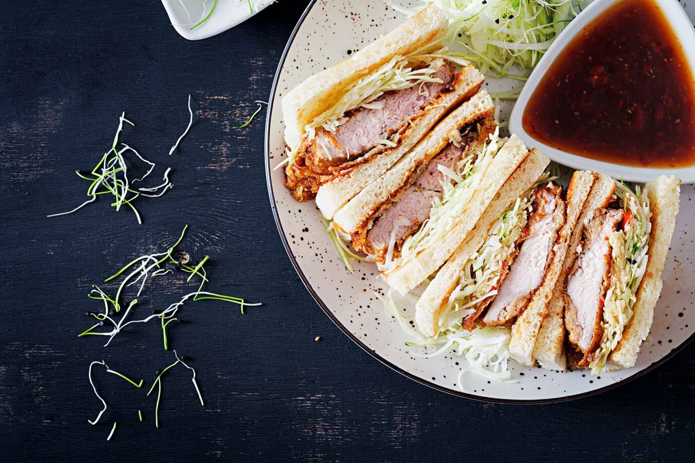 Three layers of pork cutlet sandwiches, garnished with shredded cabbage, served with a side of dipping sauce on a speckled plate.