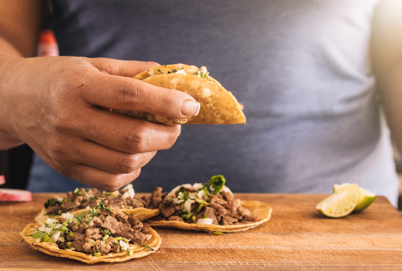 A hand holding a taco filled with meat and toppings, positioned above two tacos on a wooden table, with a lime slice beside them.