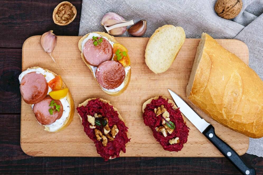 A wooden board with two open sandwiches topped with sausage, cheese, and colorful veggies, alongside a loaf of fresh bread and garlic.