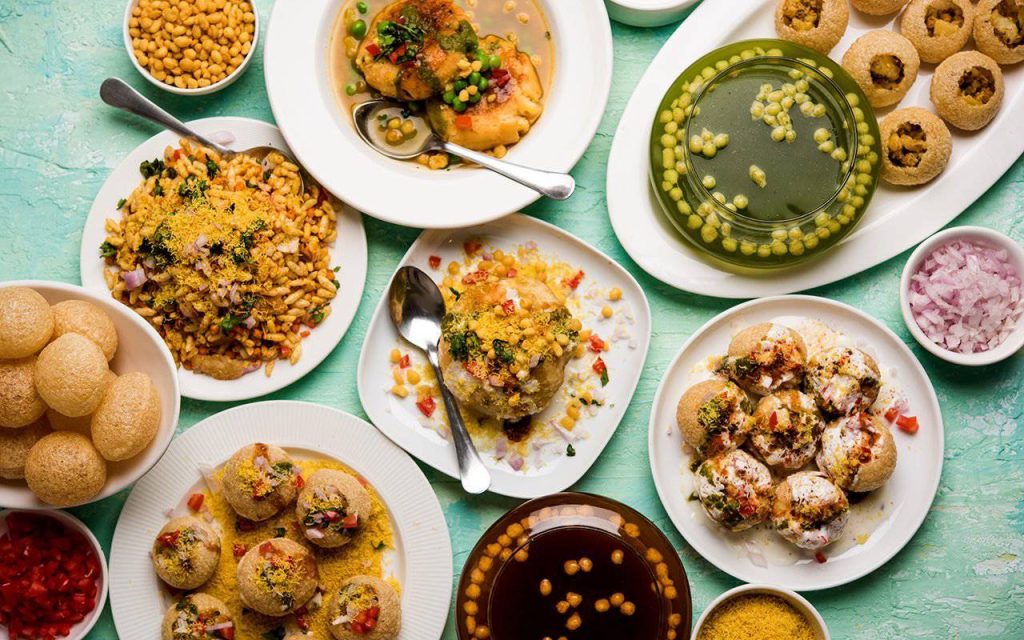 A vibrant assortment of Indian snacks including pani puri, bhel puri, and sweet treats, arranged on colorful plates and bowls.
