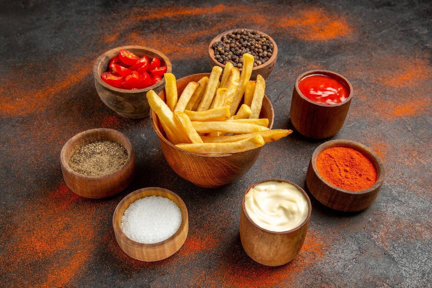 A bowl of golden French fries surrounded by wooden bowls of spices, sauces, and tomatoes on a textured, dark background.