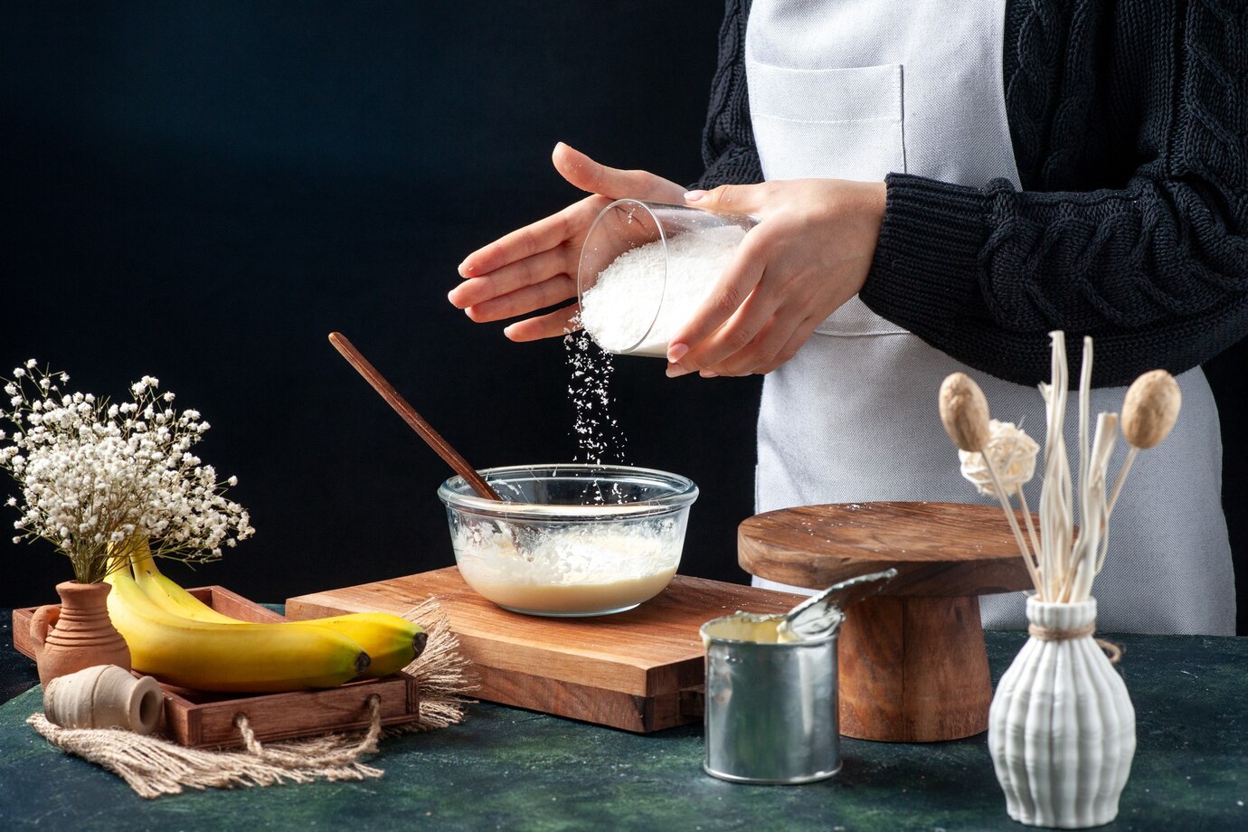 A person in a black sweater and apron pours sugar into a glass bowl, surrounded by bananas, flowers, and baking tools.