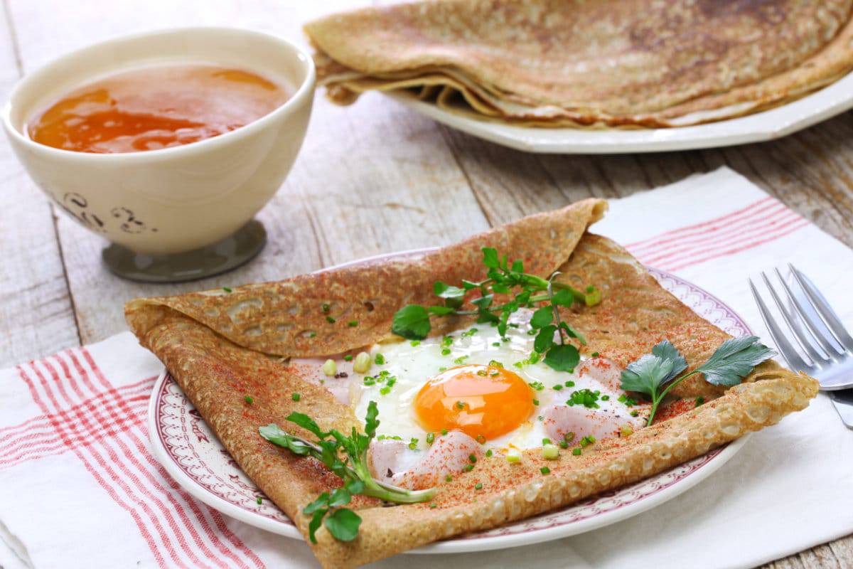 A savory buckwheat galette topped with an egg, herbs, and ham, served alongside a bowl of amber-colored drink on a rustic table.