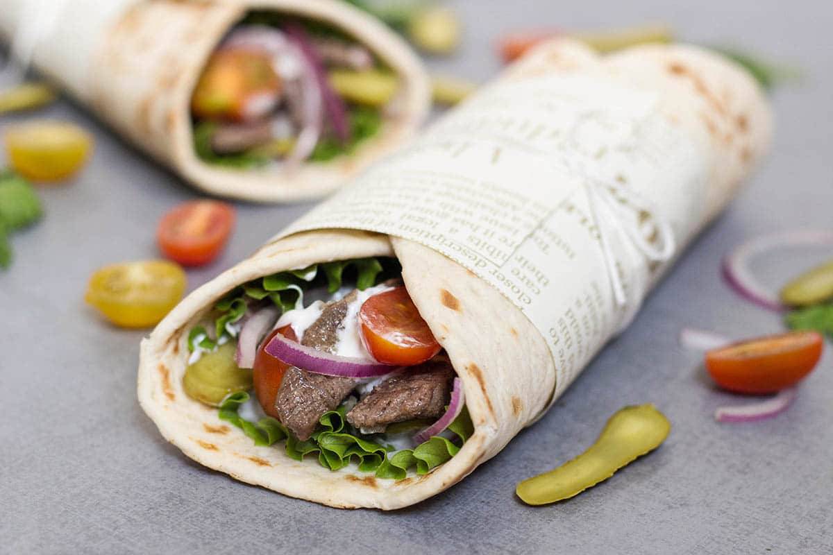 A close-up of a wrap filled with lettuce, tomatoes, pickles, onions, and meat, presented on a grey surface with scattered cherry tomatoes.