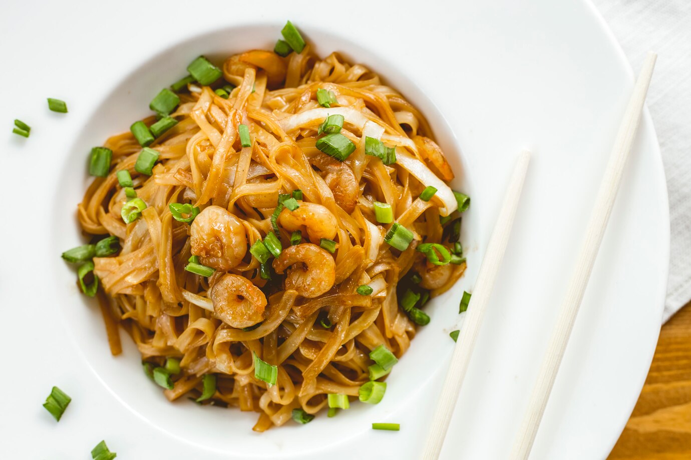 A bowl of shrimp stir-fried rice noodles garnished with green onions, accompanied by chopsticks on a wooden surface.