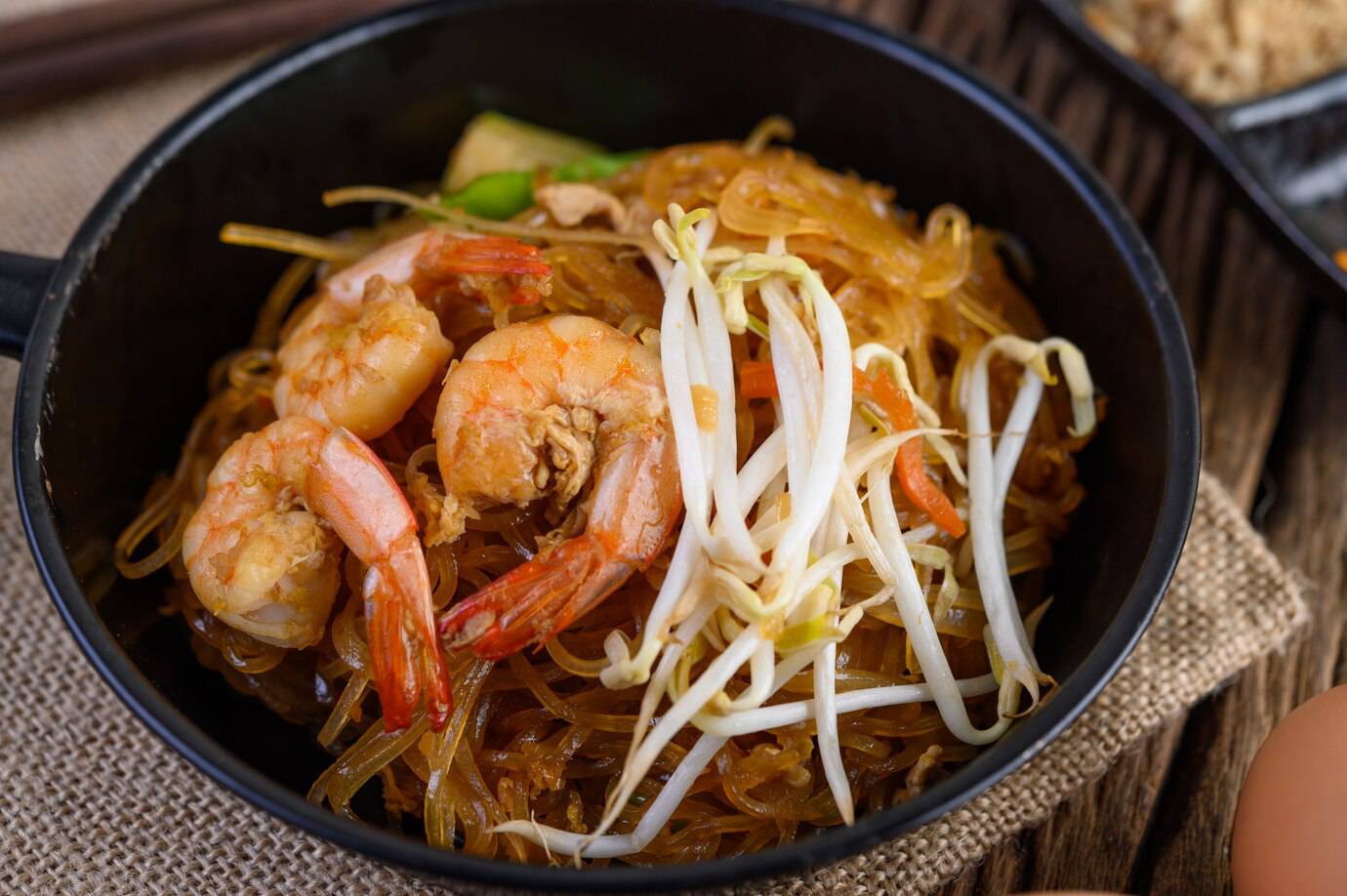 A bowl of stir-fried glass noodles topped with shrimp and bean sprouts, set on a rustic wooden background.