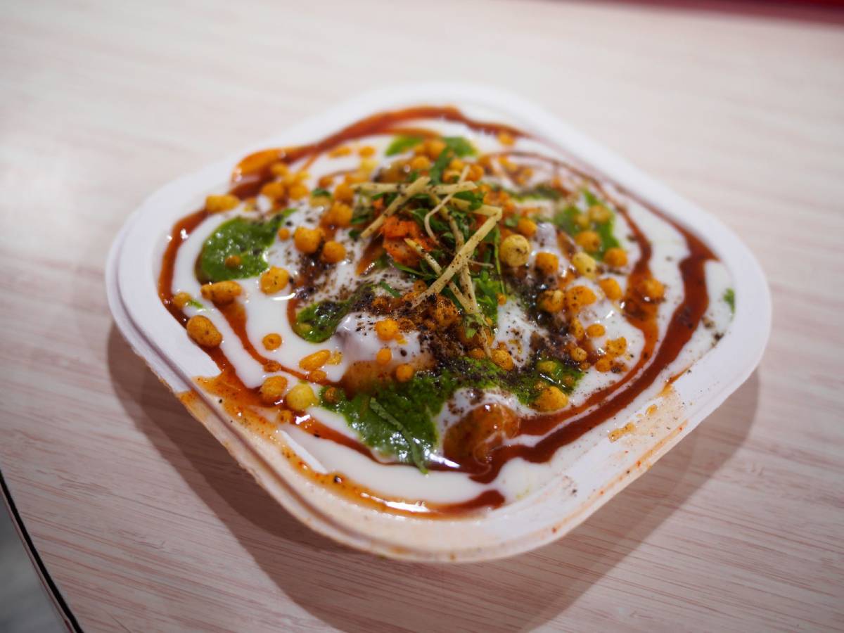 A colorful plate of dahi puri topped with yogurt, tamarind sauce, green chutney, crispy sev, and garnished with herbs.