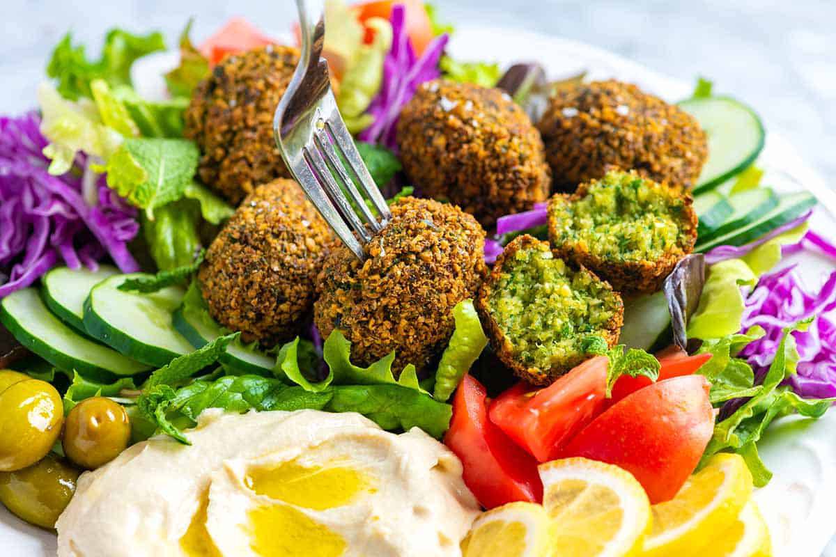 A plate of falafel balls on a bed of fresh greens, cucumbers, and tomatoes, served with hummus and lemon slices, with a fork poised.