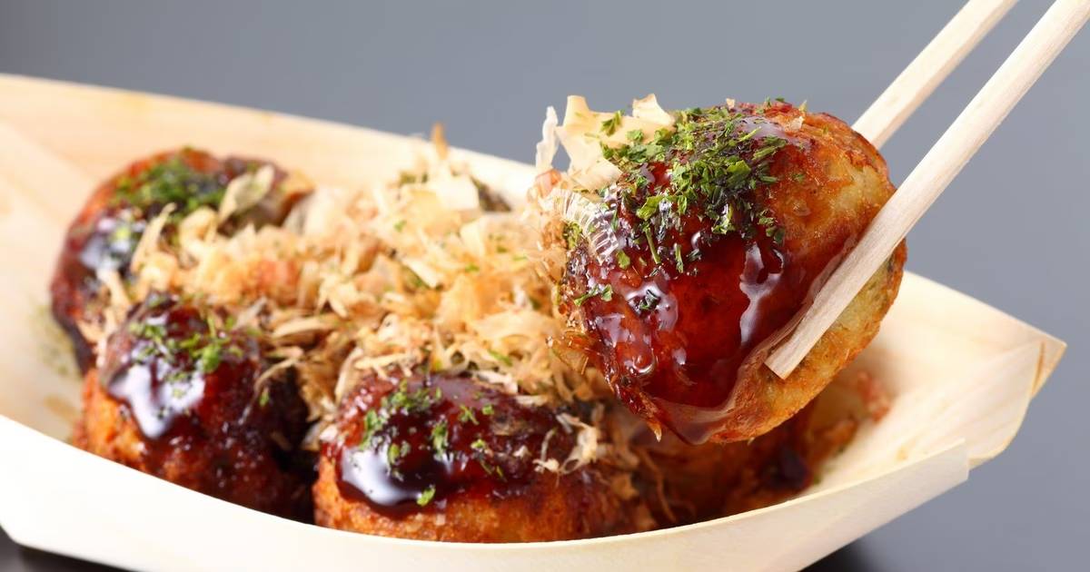 A hand holding chopsticks with a piece of takoyaki, topped with sauce, bonito flakes, and green seasoning, served in a wooden tray.