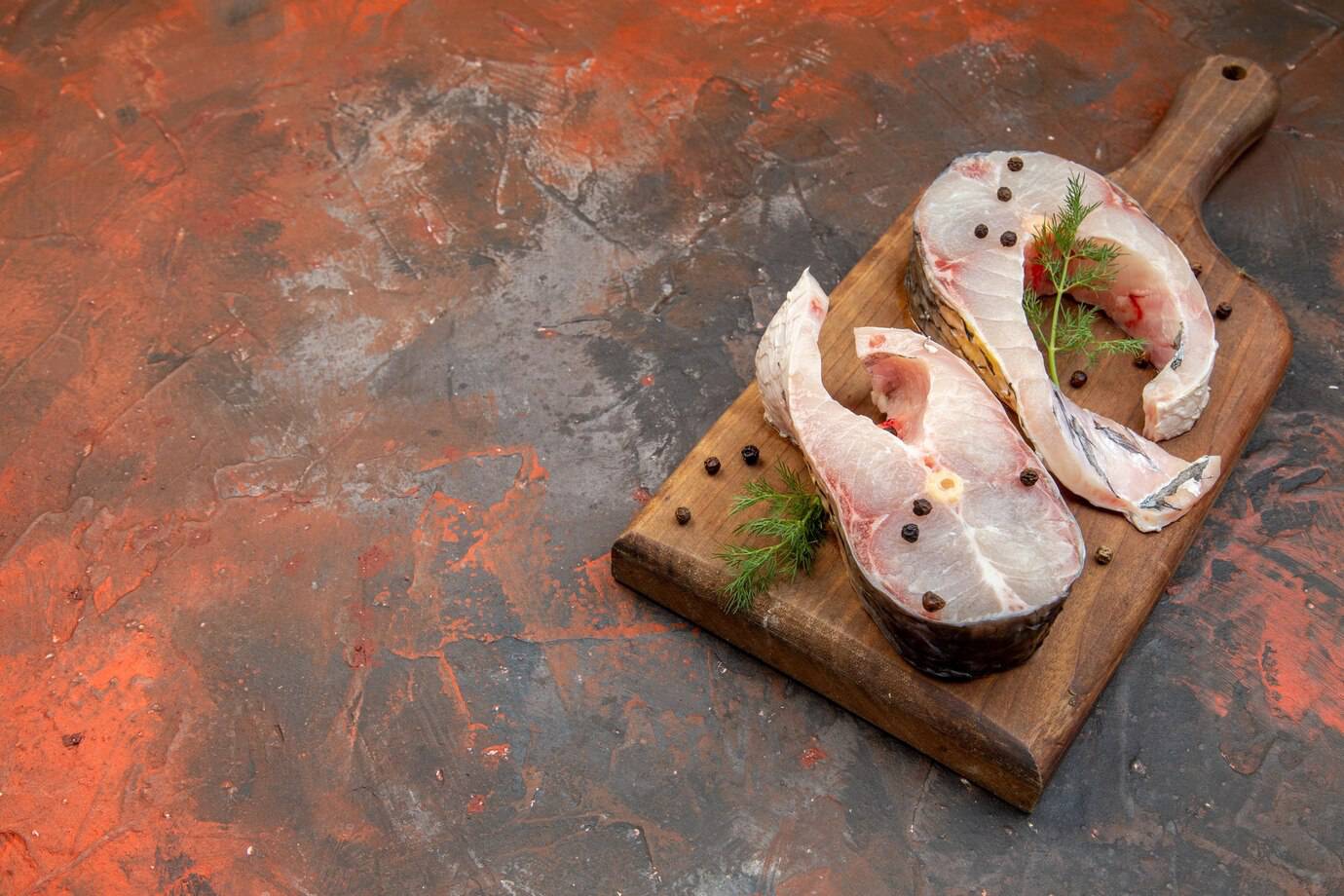 Two slices of fresh fish on a rustic wooden cutting board, garnished with dill and peppercorns against a textured orange and gray background.