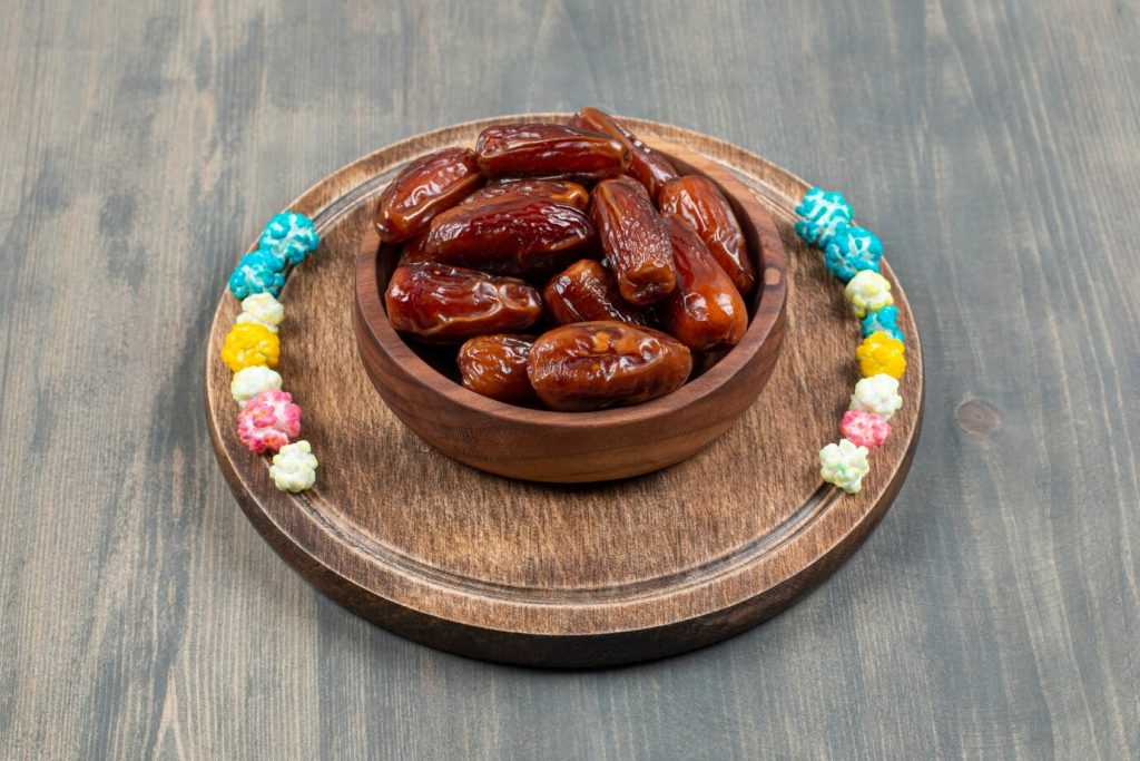 A wooden bowl filled with glossy dates sits on a wooden platter, accompanied by a colorful popcorn garland.
