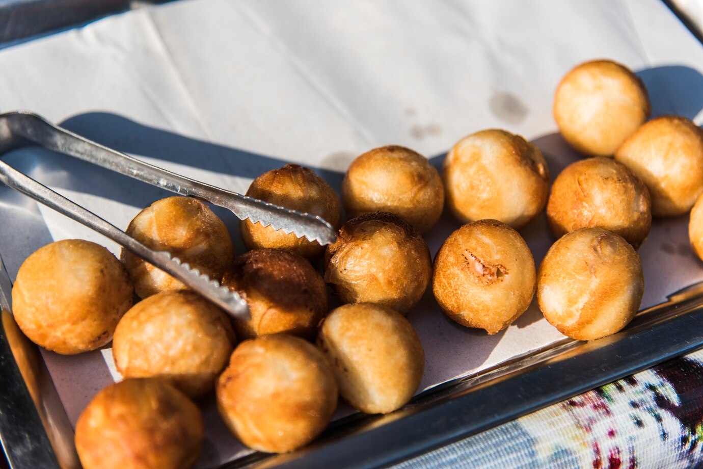 A tray filled with golden-brown fried dough balls, with tongs resting on top, set on a lightly textured tablecloth.