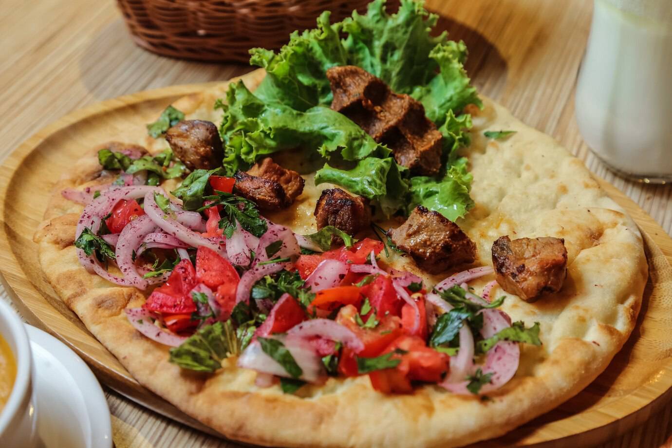 Delicious flatbread topped with grilled meat, fresh lettuce, tomato, and onion salad, served on a wooden platter.