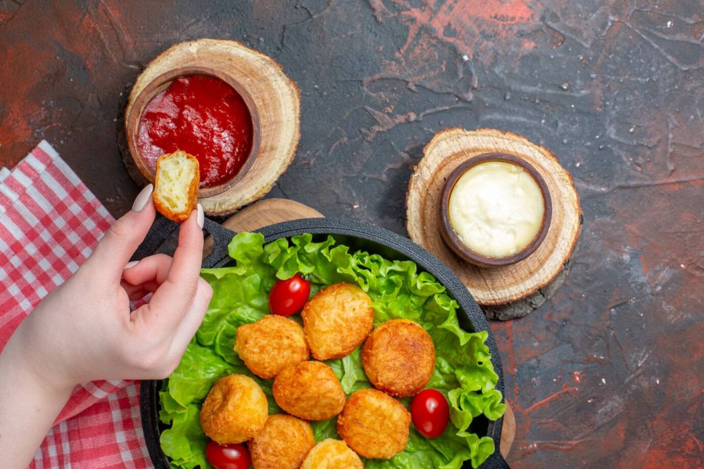 A hand holds a fried nugget above a plate of lettuce and tomatoes, with dipping sauces in rustic bowls nearby.