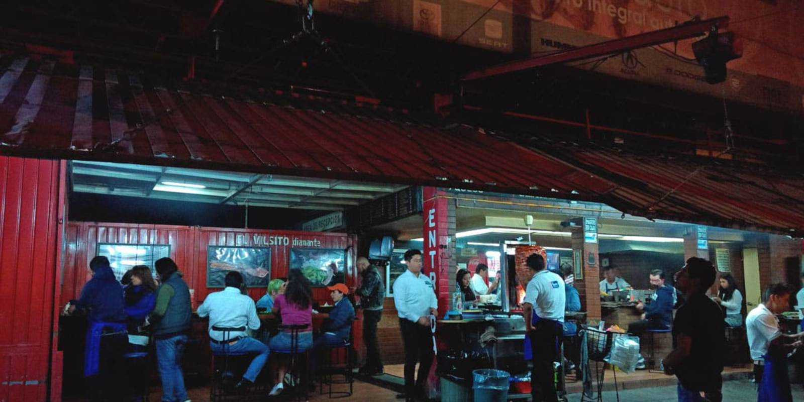 An outdoor restaurant bustling with patrons, featuring red walls, a metal roof, and staff serving food in a lively, nighttime atmosphere.
