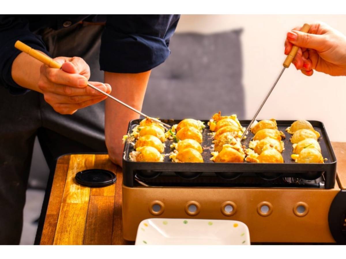 Two hands use skewers to turn golden takoyaki balls on a hot grill, with a plate ready nearby.