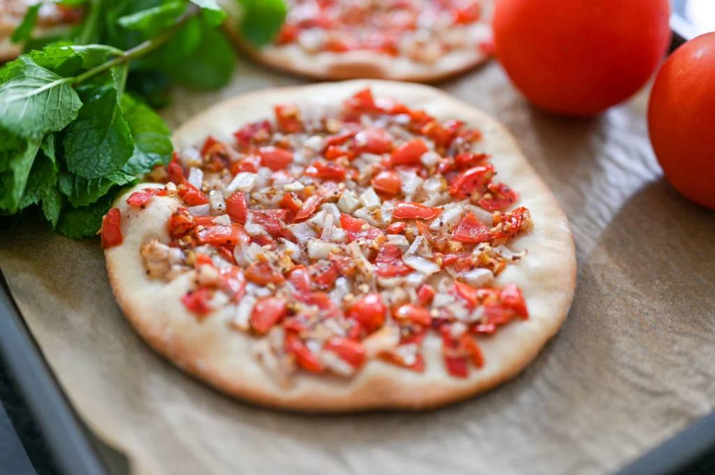 A delicious flatbread topped with diced red peppers and onions, accompanied by fresh mint leaves and ripe tomatoes.
