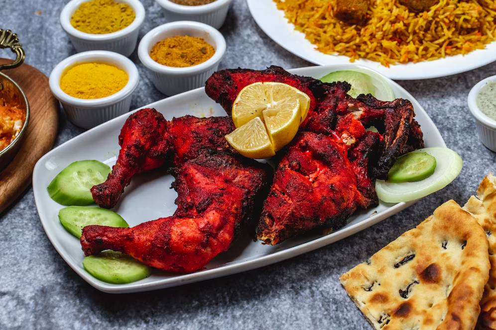 A platter of vibrant red tandoori chicken garnished with lemon and cucumber, served alongside rice, naan, and spices.