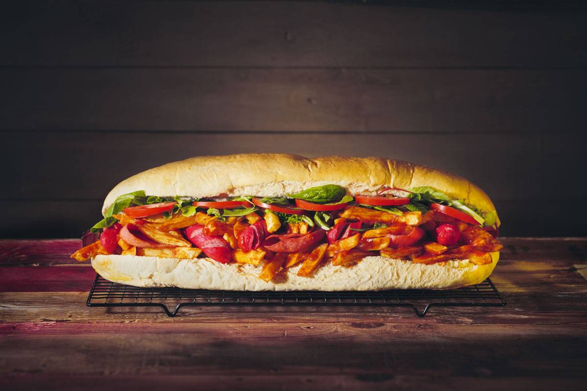 A large sub sandwich loaded with colorful tomatoes, lettuce, hot peppers, and French fries on a black cooling rack against a wooden backdrop.
