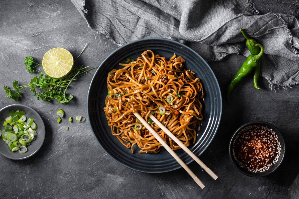 A bowl of stir-fried noodles garnished with green onions, alongside lime, coriander, chili peppers, and a dipping sauce.