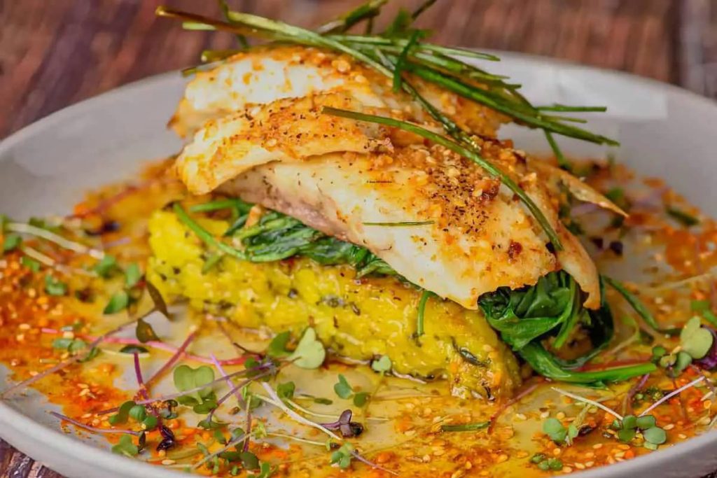 A plated dish featuring seasoned fish fillets atop spinach and yellow rice, garnished with herbs and a flavorful sauce.