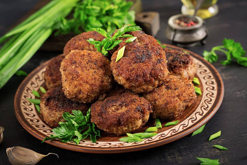 A close-up of a plate of golden-brown meat patties garnished with fresh parsley and green onions, surrounded by cloves of garlic.