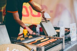 A vendor prepares hot dogs on a grill with mustard and sauces, surrounded by a colorful food stall backdrop.
