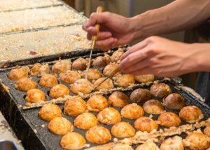 A chef uses chopsticks to expertly flip and serve golden-brown takoyaki balls on a hot grill.