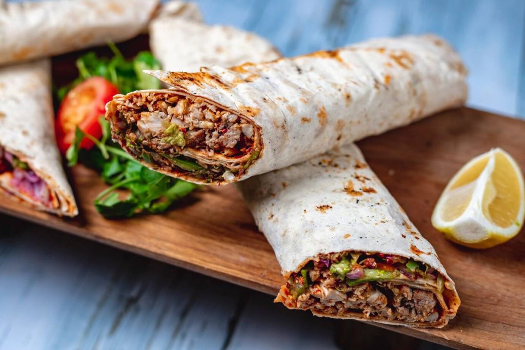 A close-up of a grilled wrap filled with meat, vegetables, and sauce, served with fresh greens and a lemon wedge on a wooden platter.