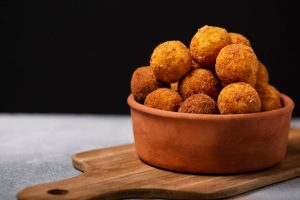 A bowl of golden crispy cheese balls stacked on a wooden board, set against a dark background.